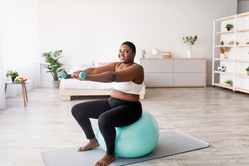 Fototapeta premium Plus size black woman working out with dumbbells on fitness ball at home, empty space. Curvy African American lady losing weight, having domestic training, leading healthy lifesyle, indoors