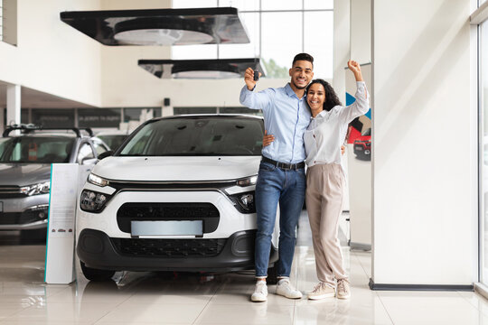 Joyful middle-eastern couple standing next to luxury white car and hugging at dealership automobile salon, rasising hands up, showing key from their new auto, cheerfully smiling at camera, full length