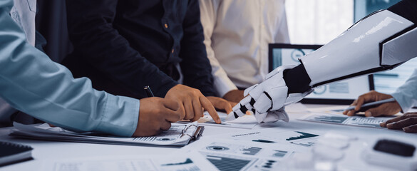 A human team collaborates with a robotic arm in an office setting, focusing on business analysis and innovative solutions for ongoing and future projects.Noogenesis