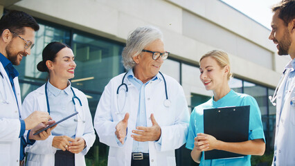 Group of Caucasian doctors gathered outside clinic. Male specialist explaining case details. Female interns reacting with smiles. Senior physician listening during training session outdoors. Teamwork.