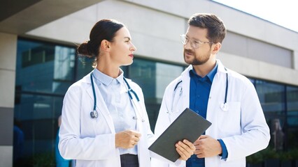 Caucasian female doctor standing outside clinic. Specialist discussing case details with male colleague holding tablet. Medical team focusing on shared evaluation. Outdoors near hospital building.