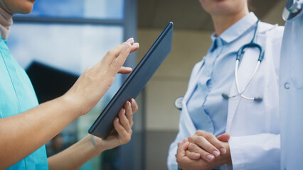 Side view showing tablet device. Female nurse tapping on screen with fingers. Two doctors discussing treatment plan calmly in front. Standing outside large hospital area. Modern technology concept.