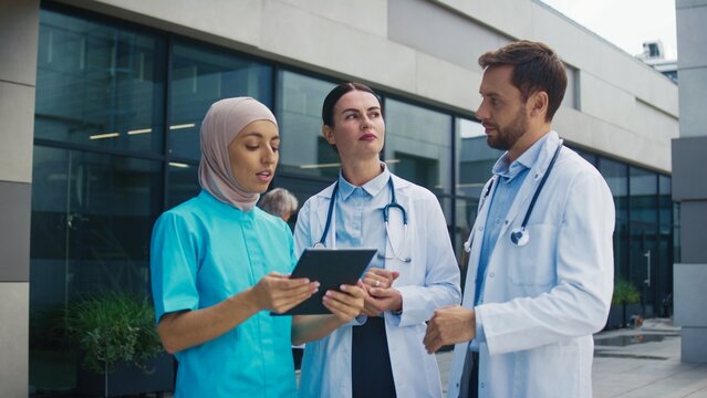 Doctors standing outside hospital. Male and female specialists listening to nurse presenting data on tablet. Team reviewing patient information calmly. Clinical setting supporting discussion. - Powered by Adobe