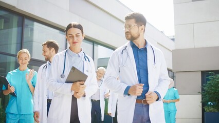 Focused medical team walking outside clinic while preparing for morning meeting. Confident female doctor holding tablet and discussing tasks with male colleague. Professionals moving together.