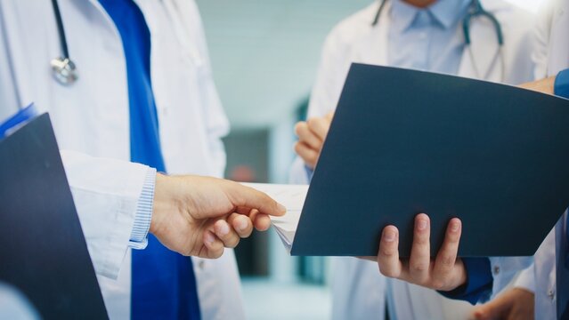 Close view of doctors exchanging papers in bright hospital hall. Reviewing lab findings. Pointing at data and clarifying details. Coordinating patient care while preparing next steps together. - Powered by Adobe
