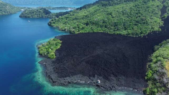 Aerial approach tilting down and turning above black volcanic lava field by coast, Banda Sea, Maluku, Indonesia.