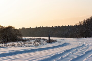 Naklejka premium Winding snowy tracks glow in the crisp winter evening. Golden hour light paints the pristine snowfield, leading toward a tranquil forest under a soft, amber sky