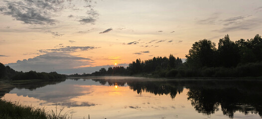 Obraz premium Sunset over calm river with trees and clouds reflecting in water at evening time