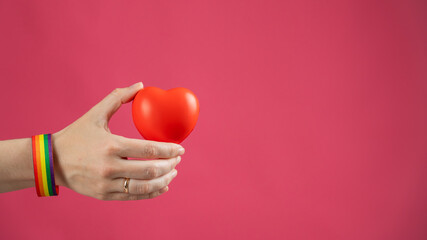 Woman with engagement ring and rainbow bracelet holding heart on pink background. Copy space. 