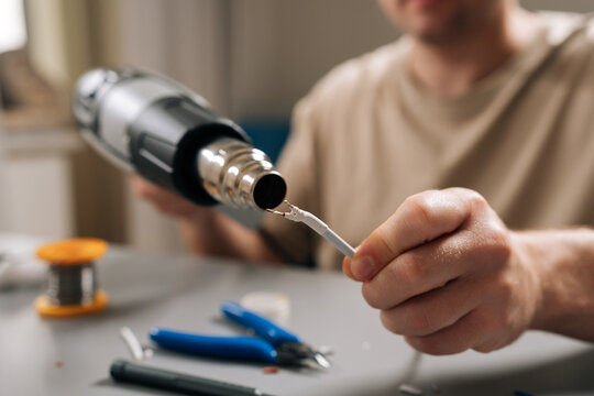 Close-up of person hands working on damaged electronic cable, applying heat with heat gun to shrink tubing or solder connecting wires, signifying diligent repair work and technical expertise.
