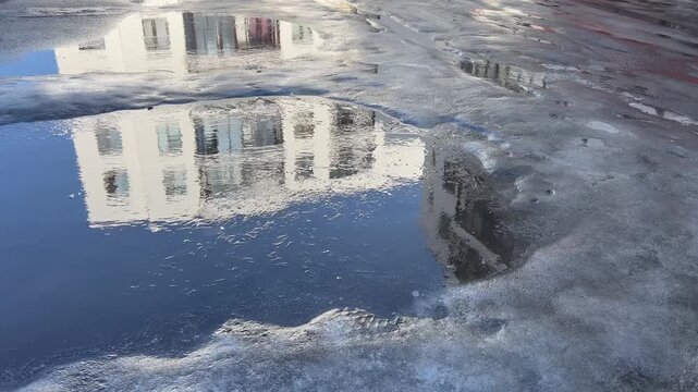 Large puddle on asphalt reflecting a modern white residential complex during spring melting and thaw