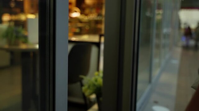 Close-up of glass storefront door handles as a hand reaches to open the entrance, revealing a warmly lit cafe interior with pastries, seating, reflections, and retail ambiance