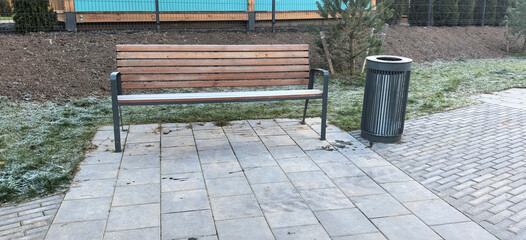 Wooden bench next to trash can in a park area with grass and stone pavement in winter time