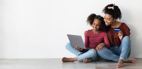 Cheerful afro-american mother and teen kid daughter buying something on Internet, using modern...