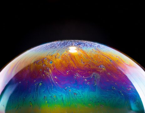 close up of a large iridescent soap bubble resting on a reflective surface with vibrant rainbow colors glowing against a black background