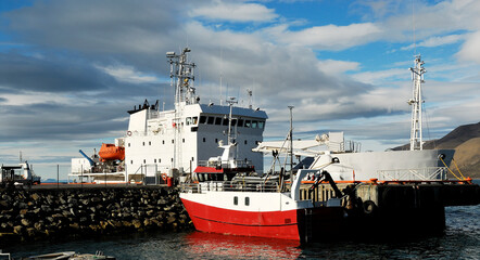 Naklejka premium Arctic Circle harbor featuring a large industrial research vessel and a smaller red and white fishing boat docked by a rocky breakwater near Greenland
