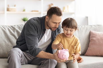 Early financial education. Cute little boy putting coin into pink piggy bank, careful daddy...
