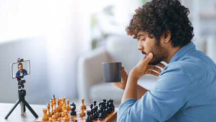 A person is seated in a living room, focused on a chess game while sipping from a cup. A smartphone on a stand shows a video call. The atmosphere is casual and engaging.