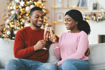 A couple sits together on a couch in a living room decorated for the holidays. They smile as they...