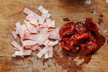 Chopped raw bacon or ham and sun dried tomatoes in oil on rustic wooden cutting board. Pasta ingredients.