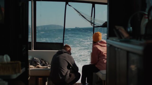 Man and woman sit on stern of yacht watching turbulent wake left by motor. View of bay surrounded by steep cliffs. Back view. Slow motion