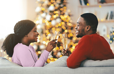Obraz premium A couple sits on a couch and toasts with glasses of sparkling drink. They are smiling and enjoying a festive atmosphere near a decorated Christmas tree with lights.