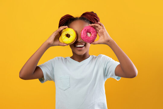 Funny afro american teen girl in white t-shirt holding colorful delicious donuts over eyes, cheerful black girl having fun while enjoying tasty sweets over yellow studio background