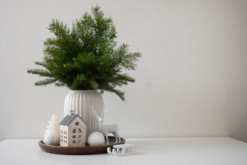 Fototapeta premium Minimal winter home decor with fir branches, ceramic house and deer figurine arranged on white table against neutral background.