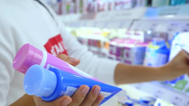 Woman holding two cosmetic tubes while another person in the background is choosing skincare products from a shelf, comparing different options and reading the labels in a supermarket