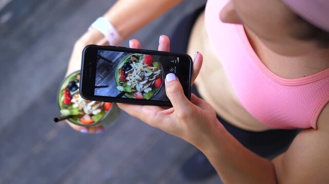 Sporty young woman taking a picture of her healthy green smoothie bowl for social media, capturing a top down view of the colorful fresh fruit, seeds, and nuts with her smartphone