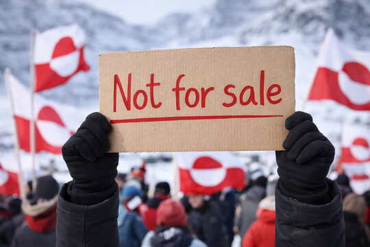 A close-up of a hand holding a cardboard sign reading "Not for sale" against a backdrop of protesters waving Greenlandic flags. Greenland's protest against American policy.