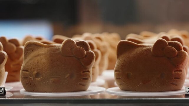 Rows of adorable cat-shaped sponge cakes with bow details on white plates in a bakery showcase, highlighting golden-brown texture and playful cartoon faces in a close-up view.