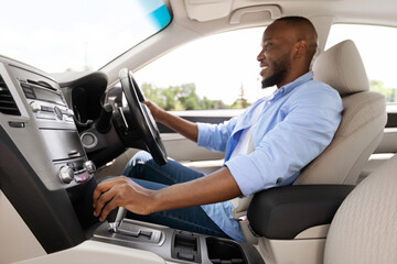 Profile side view of young African American man sitting in modern vehicle, driving his new auto, experienced male driver holding steering wheel and gear shift looking at road, selective focus on hand