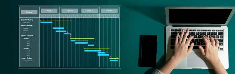 A business professional focuses on a laptop while analyzing a project management Gantt chart. This...