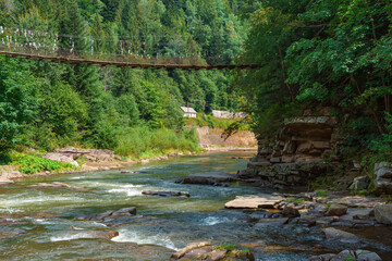 Scenic rapid mountain river, fast water flowing through dense green forest with wooden suspension bridge above. Peaceful summer landscape in Carpathian Mountains, Ukraine, travel and nature concepts.