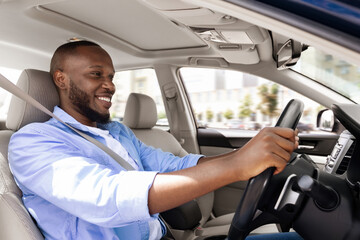 Confident Professional Driver. Side view profile portrait of smiling young African American man sitting in a car on driver's seat. Positive black guy riding in the city holding hands on steering wheel © Prostock-studio