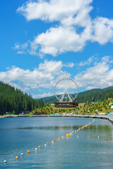 Scenic mountain resort landscape with white ferris wheel near calm lake surrounded by green forest hills. Summer travel destination, touristic recreation and leisure