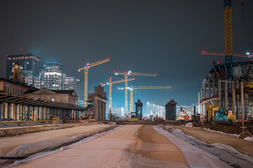 Wide night view of Ljubljana train station redevelopment: cranes tower over unfinished platforms and roadbeds, illuminated construction machinery and city skyline highlighting major urban rail