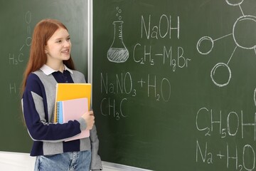 Schoolgirl with notebooks near chalkboard with chemical formulas and drawings in classroom