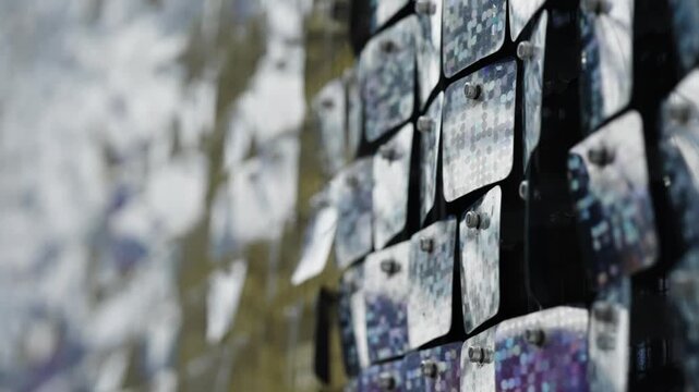 Close-up of a reflective kinetic sequin wall with shimmering square tiles, metallic mosaic panels catching light and movement, creating abstract patterns and sparkling textures