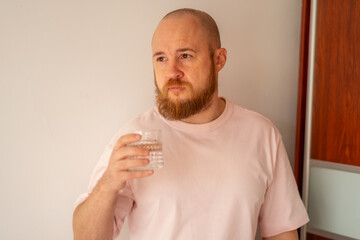 Bald man with a beard in a pink T-shirt holding a glass of water, morning routine, health biohacking concept