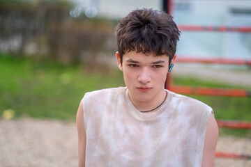 A thoughtful teenager with short dark hair rests on red outdoor bars, wearing wireless earbuds and a sleeveless top, looking calmly at the camera in an urban playground setting