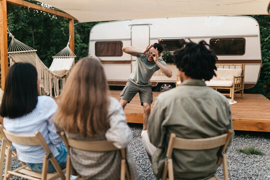 Multiracial young friends playing charades near motorhome during outdoor party. Millennial diverse people entertaining themselves with guessing game, having fun summer vacation