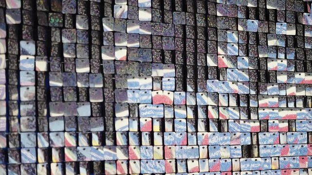 Detailed view of a kinetic mosaic mural of perforated reflective tiles, forming a colorful abstract pixelated pattern in blue, red, and white across two video frames