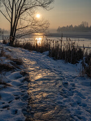 Frozen path covered with ice and snow leading toward a lake at winter sunset, golden light reflecting on the icy surface, creating a calm, serene and atmospheric winter landscape