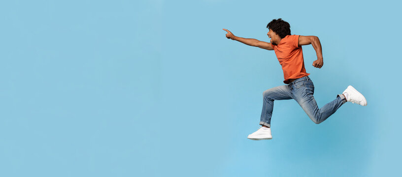Excited young black man running towards copy space, emotional african american bearded guy jumping in the air and pointing at advertisement over blue studio background, panorama, web-banner