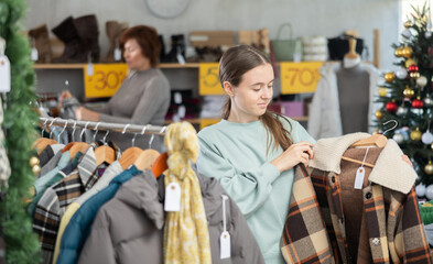 Fototapeta premium In sales area of store during Christmas sale, teen girl customer choose plaid jacket. Creating capsule wardrobe, branded clothing at affordable prices. Modern urban fashion, casual practical clothes