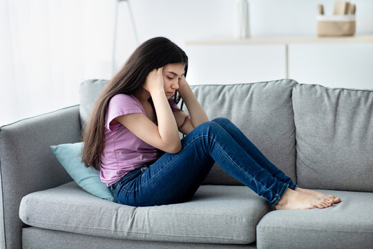 Unhappy teen girl holding her head in despair, feeling depressed, sitting on sofa at home, copy space. Indian asdolescent going through difficult times, having mental problems or mood disorder