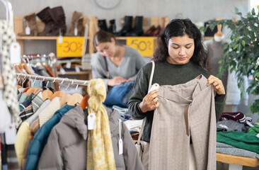 Obraz premium Latin girl is busy looking at clothes items in store, she chooses trousers. Young woman enjoys shopping for stylish winter clothes at clothing store browsing through colorful apparel on display racks