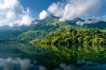 Serene verdant mountain landscape with lush forest reflected in calm lake water under partly cloudy blue sky on a bright day